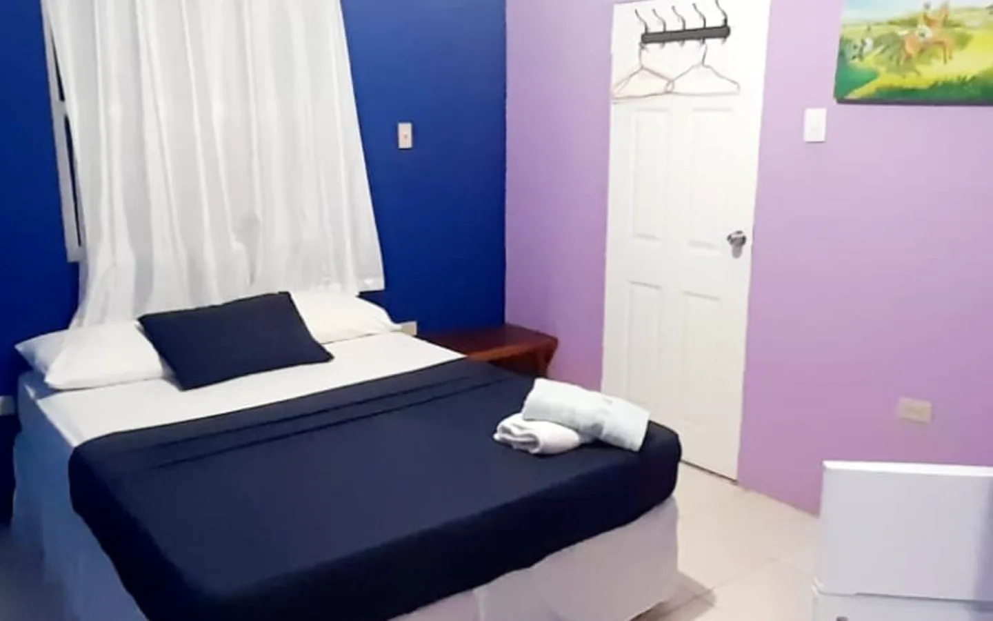 A queen bed made up in navy against a blue and lilac wall, with a coat rail on the door