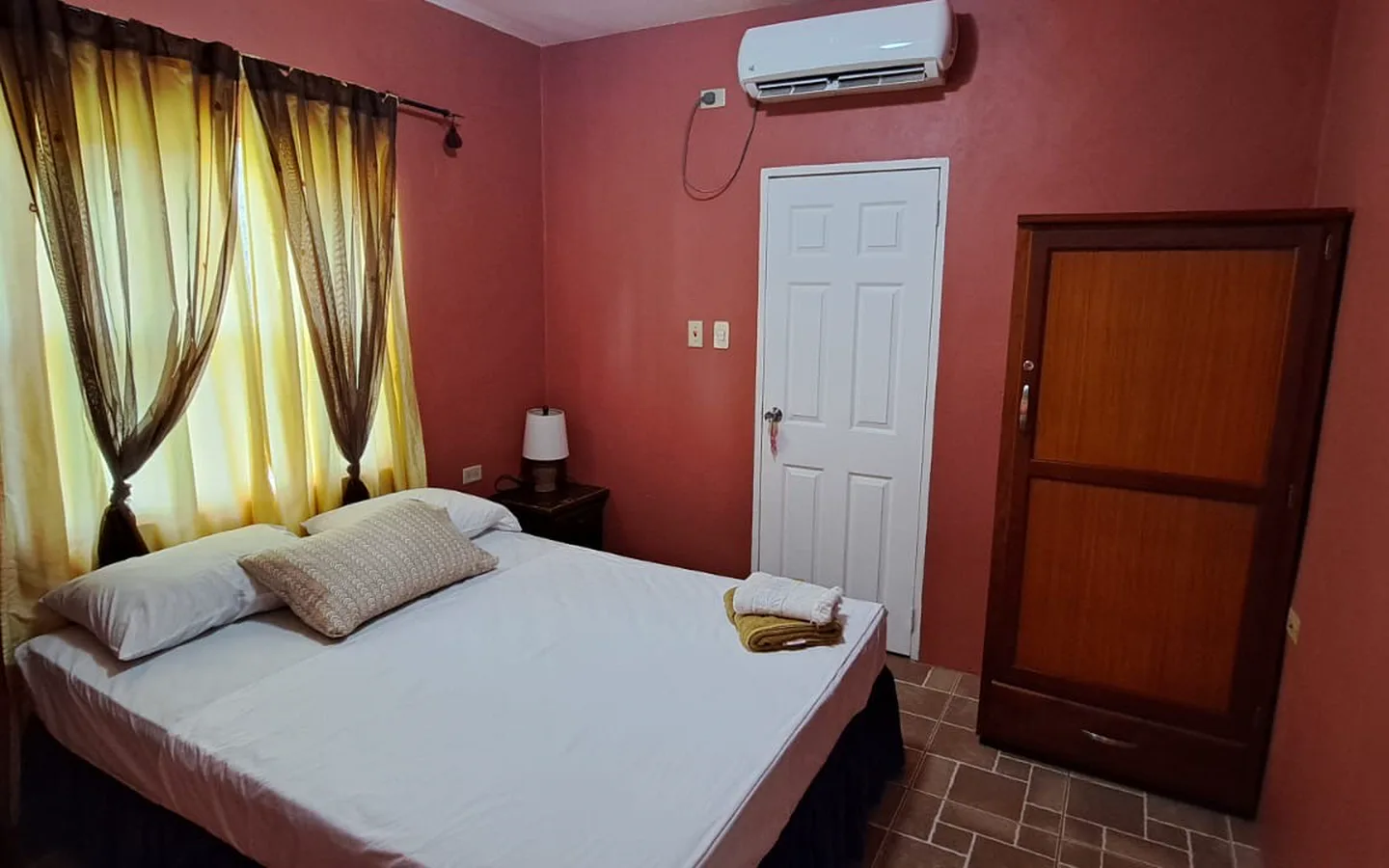 A queen bed in a terracotta room with a wardrobe and a curtained window